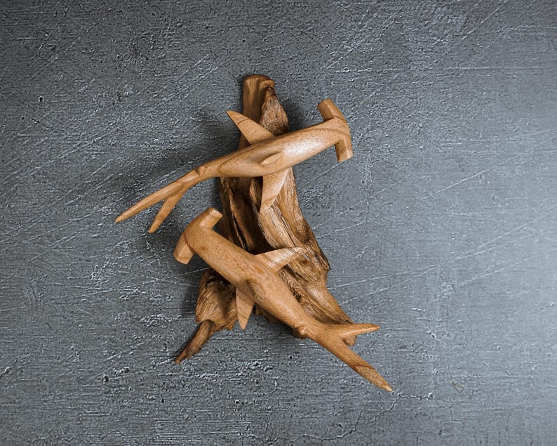 May include: Two wooden hammerhead shark sculptures are mounted on a piece of driftwood. The sharks are light brown with detailed fins and tails. The driftwood is a darker brown and has a textured surface. The background is a gray, textured surface.