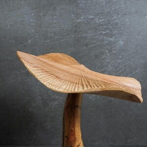 May include: A hand-carved wooden sculpture shaped like a mushroom with a wide, wavy cap featuring detailed radial grooves and a smooth, natural grain stem. The light brown wood displays organic textures and a curved, asymmetrical silhouette.