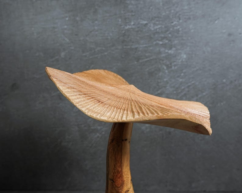 May include: A hand-carved wooden sculpture shaped like a mushroom with a wide, wavy cap featuring detailed radial grooves and a smooth, natural grain stem. The light brown wood displays organic textures and a curved, asymmetrical silhouette.