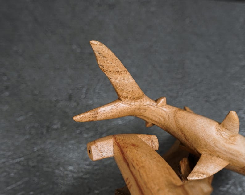 May include: Wooden shark sculptures with a natural wood grain finish. The sculptures are detailed with fins and a hammerhead shape. The sharks are set against a dark gray background.