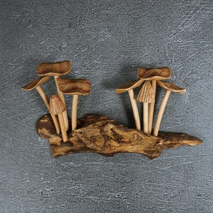May include: A decorative sculpture featuring two clusters of carved wooden mushrooms attached to a piece of textured driftwood. The mushrooms have light brown, ribbed caps and slender stems, while the base is a dark, weathered piece of wood.