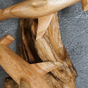 May include: Wooden sculpture featuring two stylized dolphins, carved from light-colored wood. The dolphins are attached to a textured, dark brown wooden base. The sculpture is set against a gray background.