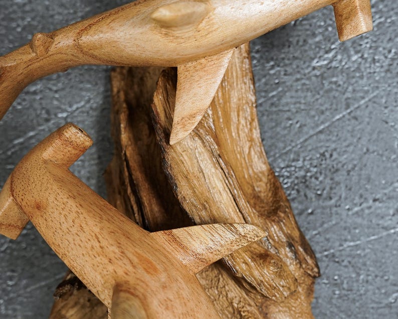May include: Wooden sculpture featuring two stylized dolphins, carved from light-colored wood. The dolphins are attached to a textured, dark brown wooden base. The sculpture is set against a gray background.