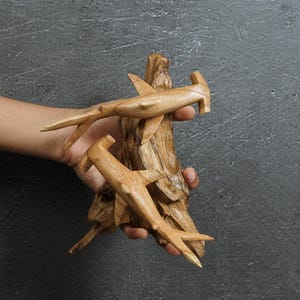 May include: Hand-carved wooden sculpture featuring two hammerhead sharks attached to a piece of driftwood. The sharks are light brown with detailed fins and tails. The driftwood has a natural, textured appearance.