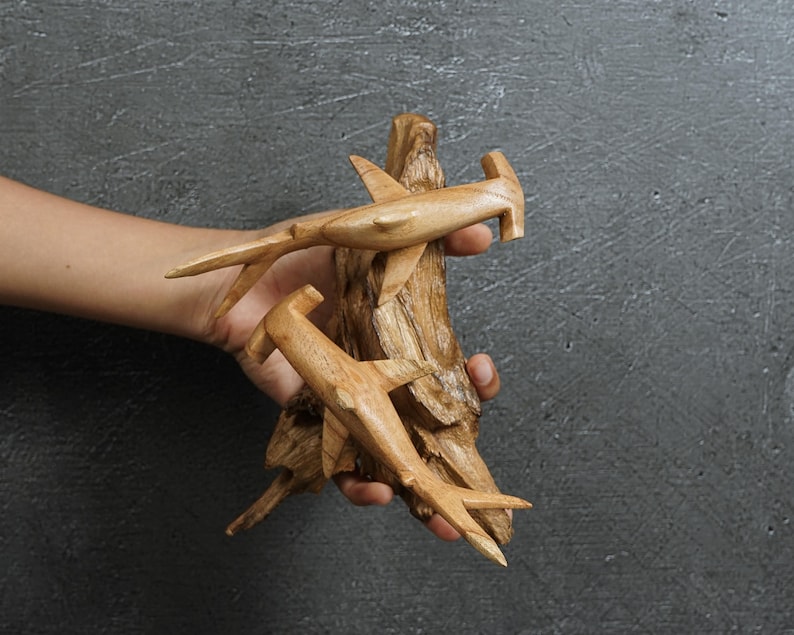 May include: Hand-carved wooden sculpture featuring two hammerhead sharks attached to a piece of driftwood. The sharks are light brown with detailed fins and tails. The driftwood has a natural, textured appearance.