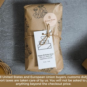 May include: A rectangular package wrapped in brown paper with a black mushroom pattern, secured with twine. A round Wood Rose Craft sticker and a white gift tag are attached. The tag features a heart icon and the text: Happy Birthday Anya.