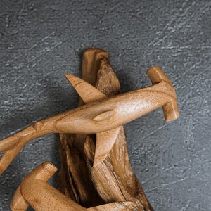 May include: Wooden hammerhead shark sculpture with detailed fins and tail, mounted on a piece of driftwood. The shark is carved from light brown wood, showcasing the natural grain. The sculpture is set against a gray textured background.
