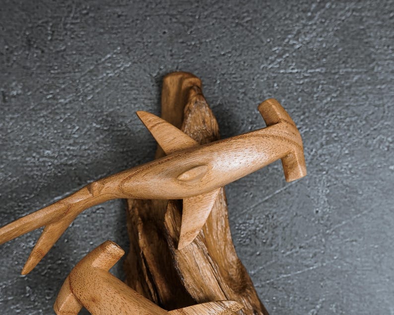May include: Wooden hammerhead shark sculpture with detailed fins and tail, mounted on a piece of driftwood. The shark is carved from light brown wood, showcasing the natural grain. The sculpture is set against a gray textured background.