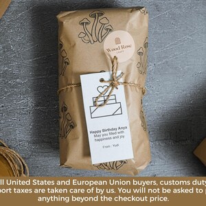 May include: A rectangular package wrapped in brown paper with a black mushroom pattern, secured with twine. A round Wood Rose Craft sticker and a white gift tag are attached. The tag features a heart icon and the text: Happy Birthday Anya.