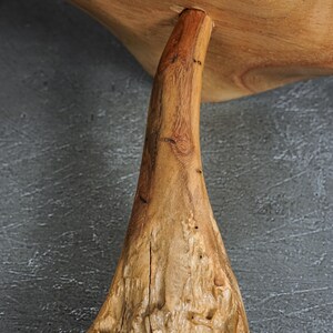 May include: A natural light brown wooden stand featuring a thick, textured, organic-shaped base that tapers upward to support a smooth, flat, circular top surface. The wood grain is visible throughout, showcasing knots and variations in the texture.