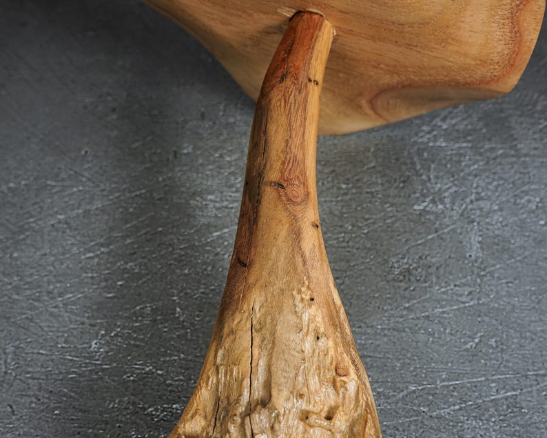 May include: A natural light brown wooden stand featuring a thick, textured, organic-shaped base that tapers upward to support a smooth, flat, circular top surface. The wood grain is visible throughout, showcasing knots and variations in the texture.