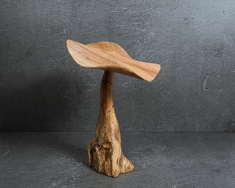 May include: A hand-carved wooden sculpture shaped like a mushroom with a wide, wavy cap and a thick, textured stalk. The wood displays natural grain patterns and light brown tones throughout the piece, showcasing the organic texture of the material.