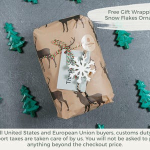 Pu&ograve; includere: Un regalo rettangolare avvolto in carta marrone con un motivo di cervi marrone scuro. Il pacchetto &egrave; legato con uno spago rosso, bianco e verde, decorato con un ornamento a forma di fiocco di neve bianco e un cartellino grigio chiaro con il testo Thank you.