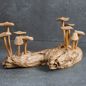 Può includere: Una scultura decorativa con molteplici funghi in legno marrone chiaro, intagliati con cappelli e gambi dettagliati, fissati su un pezzo di legno galleggiante testurizzato. I funghi variano in altezza e forma del cappello in un gruppo.