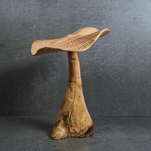 May include: A hand-carved wooden sculpture shaped like a mushroom with a wide, wavy cap featuring radial grooves and a thick, textured stem. The natural light brown wood grain is visible throughout the piece, showcasing organic knots and variations.