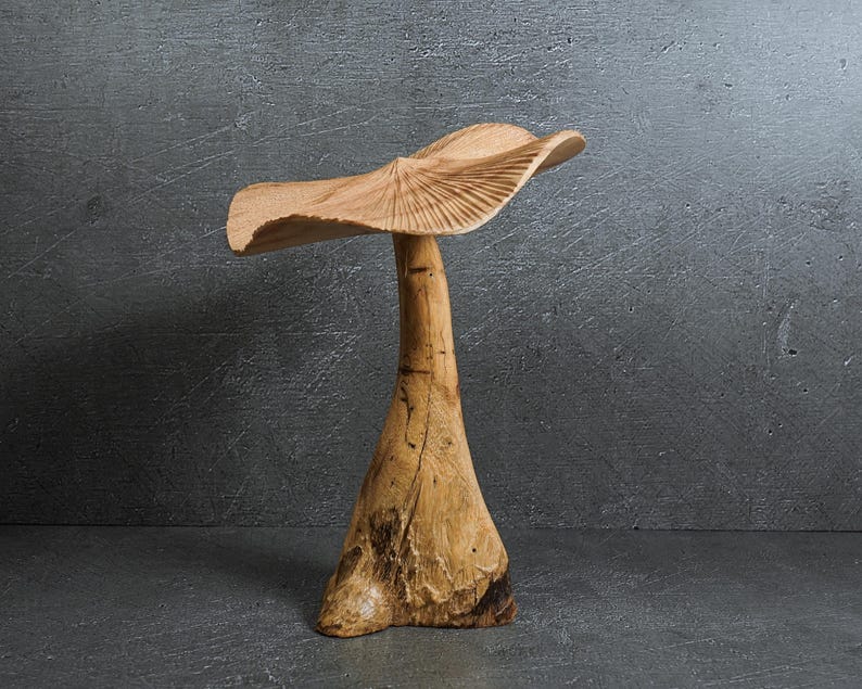 May include: A hand-carved wooden sculpture shaped like a mushroom with a wide, wavy cap featuring radial grooves and a thick, textured stem. The natural light brown wood grain is visible throughout the piece, showcasing organic knots and variations.