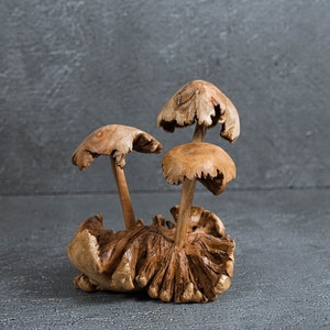 May include: A wooden sculpture of three mushrooms with brown and tan tones. The mushrooms are arranged in a cluster with the largest mushroom in the center.