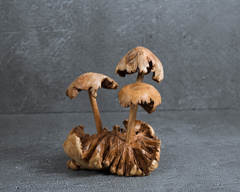 May include: A wooden sculpture of three mushrooms with brown and tan tones. The mushrooms are arranged in a cluster with the largest mushroom in the center.