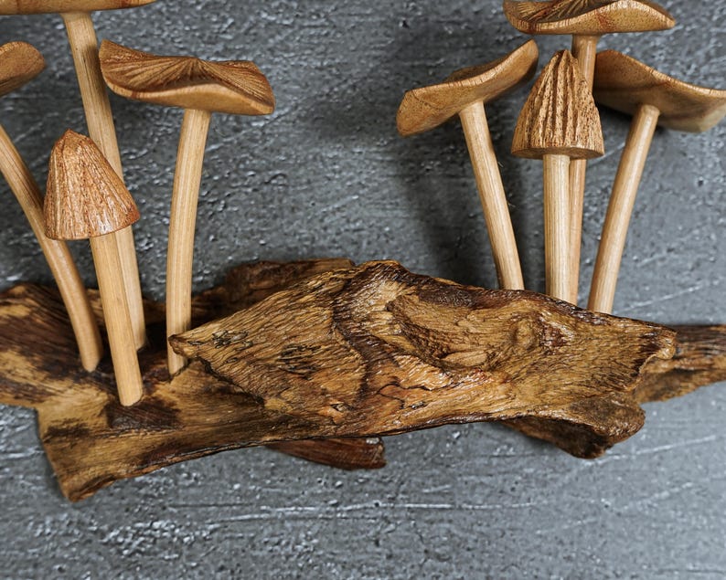 May include: A decorative sculpture featuring multiple carved wooden mushrooms with smooth, light brown stems and textured caps, all attached to a piece of dark, weathered natural driftwood with a prominent grain and irregular, organic shape.