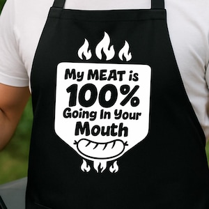 May include: Black apron with the text "My MEAT is 100% Going In Your Mouth" in white. A person is wearing the apron while grilling sausages on a barbecue. The person is smiling and holding tongs.