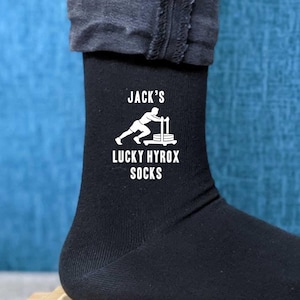 May include: Black athletic socks with the text "Jack's Lucky Hyrox Socks" in white. The socks feature a graphic of a weightlifter. The socks are on a wooden surface.