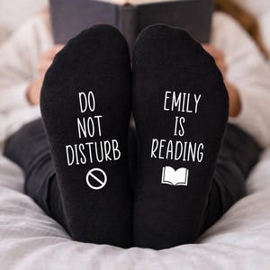 May include: Black socks with white text: "DO NOT DISTURB" with a no symbol and "EMILY IS READING" with a book icon. A person is wearing the socks while reading a book.