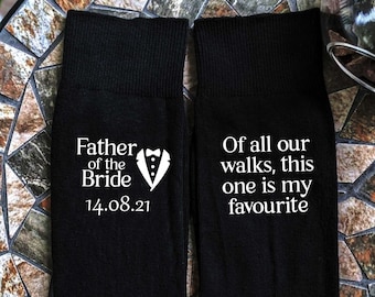May include: A pair of black socks with white text. One sock says "Father of the Bride 14.08.21" and the other sock says "Of all our walks, this one is my favourite".