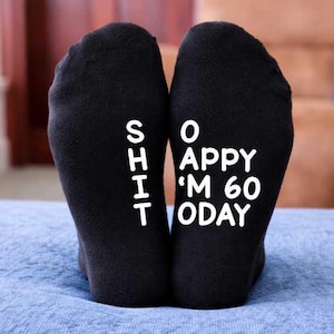 May include: A pair of black socks with the words "SHIT I'M 60 TODAY" and "SO HAPPY" printed in white. The socks are on a blue cushion, with a brown sofa visible in the background. A humorous birthday gift.