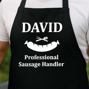 Pode incluir: Avental preto com o nome "DAVID" em branco, um gráfico de salsicha e o texto "Professional Sausage Handler". Uma pessoa sorri enquanto usa o avental perto de uma grelha com salsichas.
