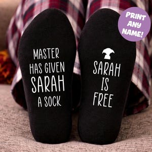 Può includere: Calzini neri con testo bianco. Uno dice "MASTER HAS GIVEN SARAH A SOCK", l'altro "SARAH IS FREE". Un cerchio viola dice "PRINT ANY NAME!"