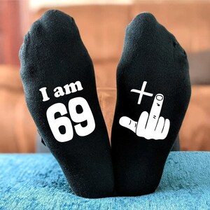 May include: A pair of black socks with white text. One sock says "I am 69" and the other sock has a white plus sign and a middle finger.