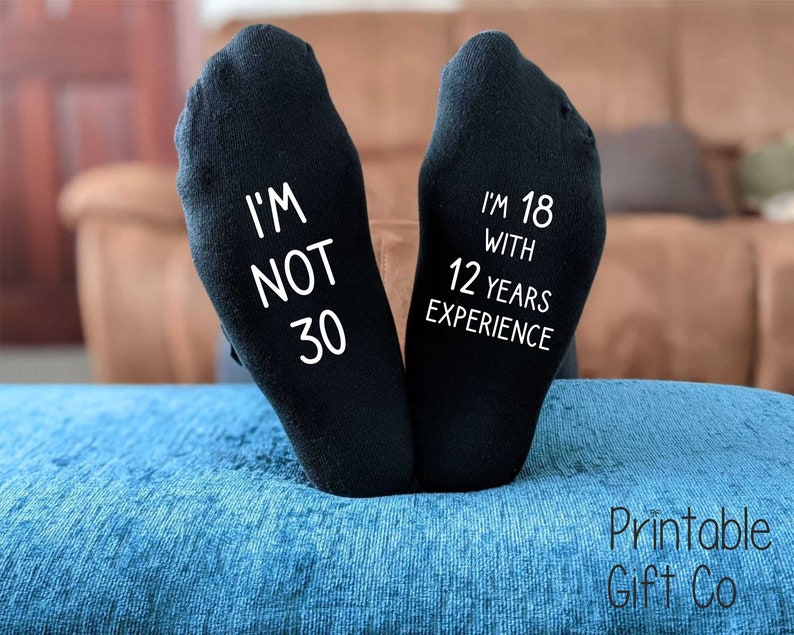 May include: A pair of black socks with white text. One sock says "I'm not 30" and the other sock says "I'm 18 with 12 years experience".