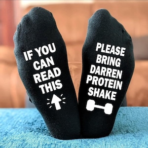 Peut inclure: Chaussettes noires avec texte blanc. L'une dit « IF YOU CAN READ THIS » avec une flèche. L'autre dit « PLEASE BRING DARREN PROTEIN SHAKE » avec un haltère. Les chaussettes sont sur une surface bleue.