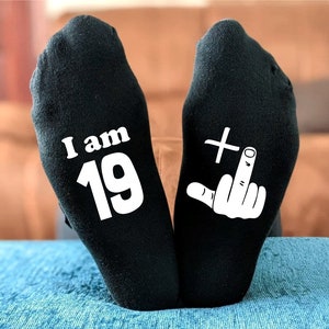 May include: A pair of black socks with white text. One sock says "I am 19" and the other sock has a white middle finger with a plus sign above it.