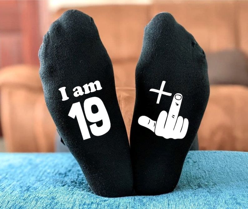 May include: A pair of black socks with white text. One sock says "I am 19" and the other sock has a white middle finger with a plus sign above it.