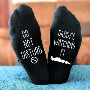 May include: Black socks with white text. One sock reads "DO NOT DISTURB" with a no symbol. The other sock says "DADDY'S WATCHING F1" with a race car graphic. The socks are on a blue surface.