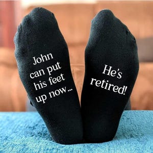 Könnte beinhalten: Schwarze Socken mit weißem Text. Auf einer Socke steht "John kann jetzt die Füße hochlegen...", auf der anderen "Er ist in Rente!". Die Socken liegen auf einer blauen Oberfläche, ein Geschenk.