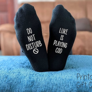 Puede incluir: Un par de calcetines negros con texto blanco. Un calcetín dice "No molestar" con un símbolo de prohibido el paso. El otro calcetín dice "Luke está jugando a COD".