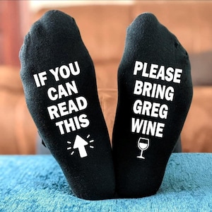 Peut inclure: Chaussettes noires avec texte blanc. L'une dit "IF YOU CAN READ THIS" avec une flèche vers le haut. L'autre dit "PLEASE BRING GREG WINE" avec un verre de vin. Un cadeau amusant et original.