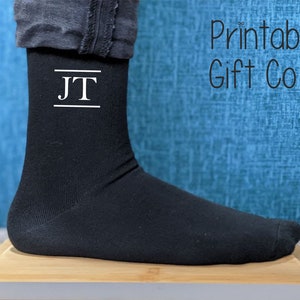 May include: Black sock with the initials "JT" in white, displayed in a classic serif font, with a line above and below the initials. The text "Printable Gift Co" is in the upper right corner. The sock is on a wooden surface.