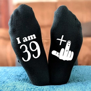 May include: A pair of black socks with white text. One sock says "I am 39" and the other sock has a white middle finger with a plus sign above it.