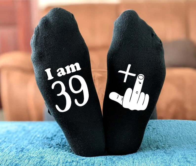 May include: A pair of black socks with white text. One sock says "I am 39" and the other sock has a white middle finger with a plus sign above it.