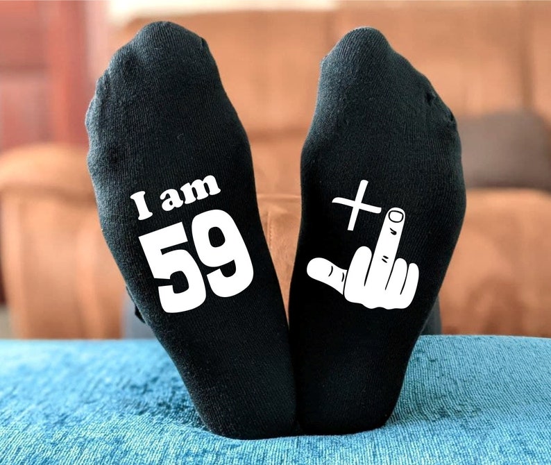 May include: A pair of black socks with white text. One sock says "I am 59" and the other sock has a white middle finger with a plus sign above it.