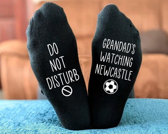 Puede incluir: Un par de calcetines negros con texto blanco. Un calcetín dice "No molestar" con una señal de prohibido el paso. El otro calcetín dice "El abuelo está viendo el Newcastle" con un balón de fútbol.