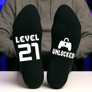 Peut inclure: Une paire de chaussettes noires avec du texte blanc. Une chaussette dit "LEVEL 21" et l'autre dit "UNLOCKED" avec une icône de manette de jeu.