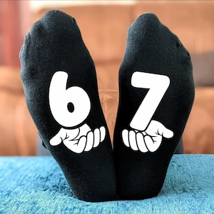 Pode incluir: Meias pretas com os números 6 e 7 impressos em branco nas solas. Cada número é acompanhado por um gráfico branco de uma mão. As meias são exibidas em uma superfície azul.
