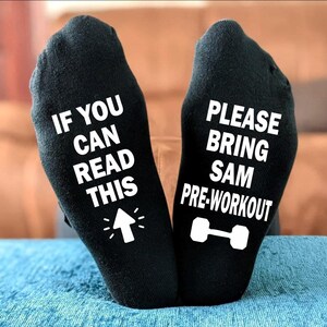 Peut inclure: Paire de chaussettes noires avec texte blanc. Une chaussette dit "IF YOU CAN READ THIS" avec une flèche. L'autre dit "PLEASE BRING SAM PRE-WORKOUT" avec un haltère. Les chaussettes sont présentées sur une surface bleue.