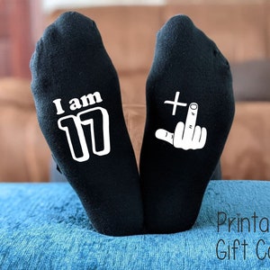 May include: A pair of black socks with white text. One sock says "I am 17" and the other sock has a white middle finger with a plus sign.