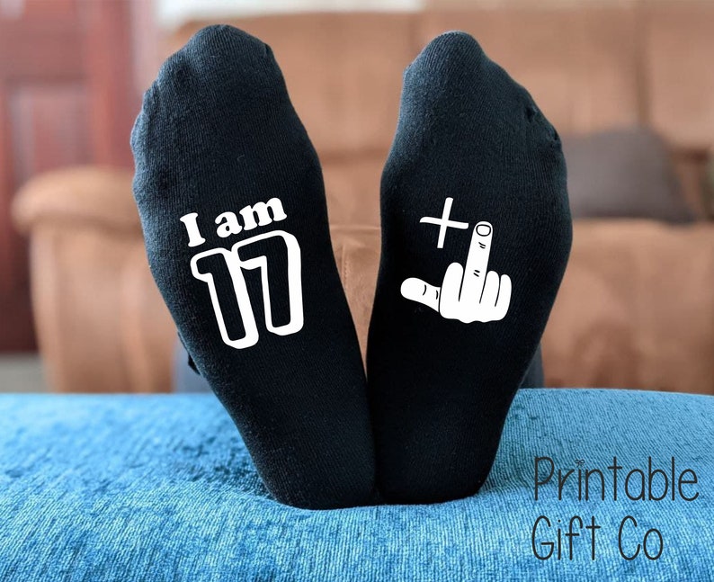 May include: A pair of black socks with white text. One sock says "I am 17" and the other sock has a white middle finger with a plus sign.