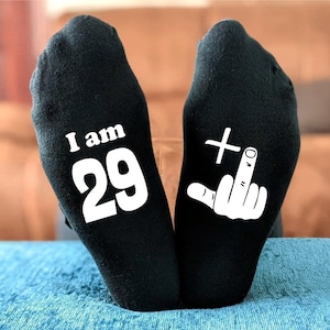 May include: A pair of black socks with white text. One sock says "I am 29" and the other sock has a white middle finger with a plus sign above it.